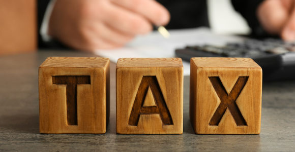 Wooden blocks spelling out the word TAX with a person writing and calculating paperwork in the background.